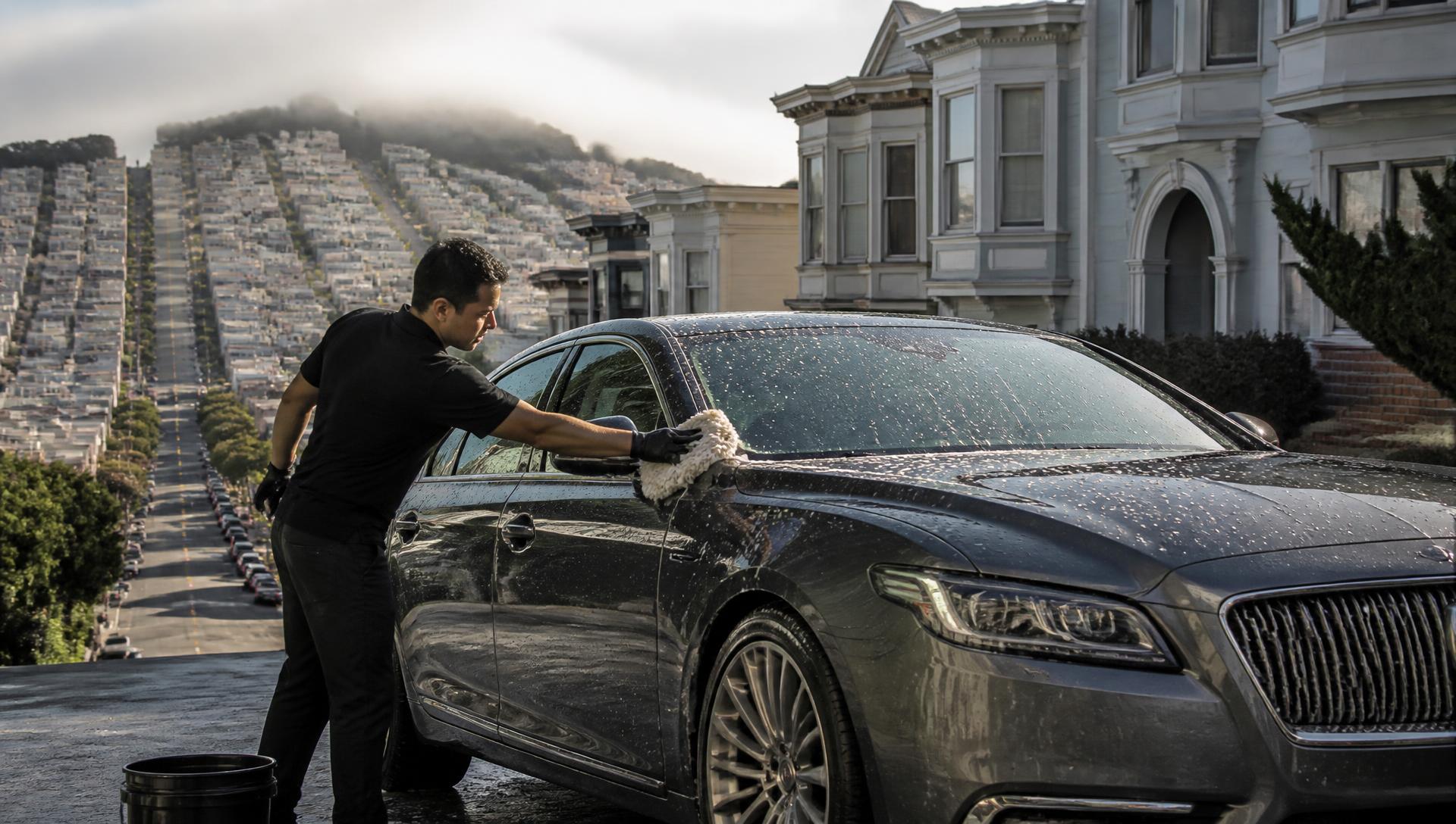 Detailer hand washing a dark grey sedan on a steep San Francisco residential street
